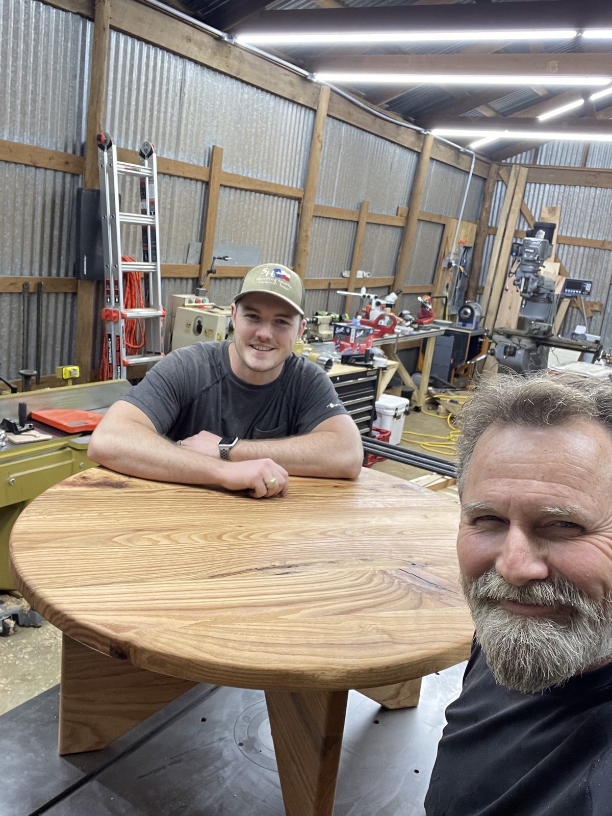Round solid-wood tabletop on a trestle base in the woodshop, two people beside it